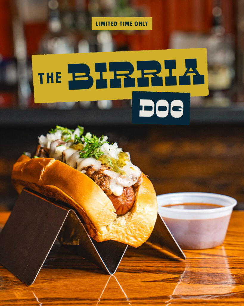 The Birria Dog: an all beef hotdog topped with house made birria, white cheddar, salsa verde, cilantro and onion, served with a side of dipping sauce
